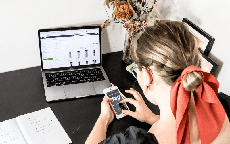 catalogo de productos: imagen de una mujer rubia trabajando con su movil con un portatil sobre la mesa y un catalogo de productos de facebook apareciendo en la pantalla