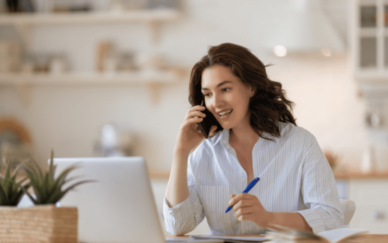 Quais São os 9 Tipos de Campanhas do Google Ads? Tipos de Campanhas Google Ads: mulher sentada em frente a um computador, com camisa branca, segurando uma caneta e um celular no ouvido.