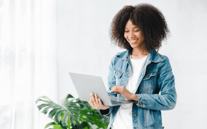 Robotic Process Automation: imagem de uma jovem negra, vestida com uma camisa branca e jaqueta jeans, em pé, segurando um laptop.