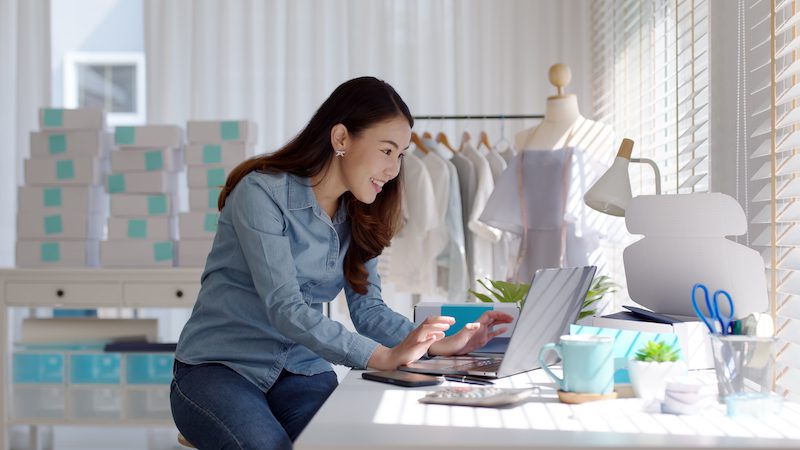 Automação para e-commerce: imagem de uma mulher asiática digitando em um notebook com caixas de encomendas ao fundo