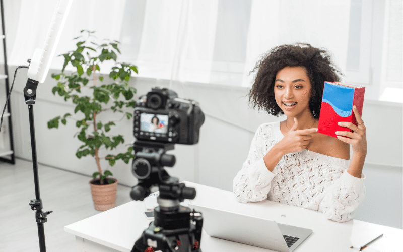 99 influencia: imagen de una cámara apuntando a una mujer negra que graba un vídeo con un libro en las manos