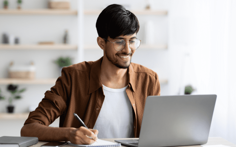 planejador de palavras chaves: imagem de um homem jovem, usando óculos, com camisa marrom, sentado, digitando em um notebook com uma mão e anotando uma informação em um papel com a outra mão