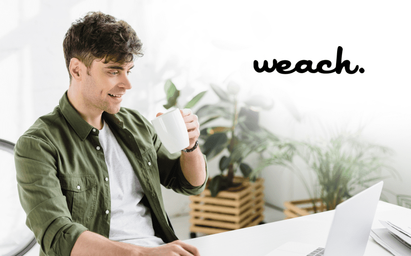 Weach group: man smiling, with a white cup in his hand, looking at his computer.