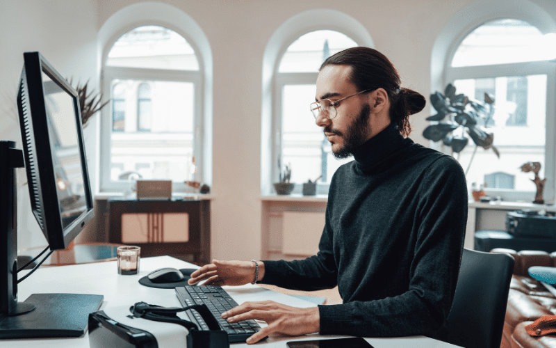 AI for Programming: young man with a beard, hair tied back, black turtleneck, wearing glasses and looking at his computer.